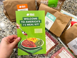 A bunch of HelloFresh bagged items laid out on the counter with a person holding up a Brochure for HelloFresh