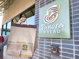 A person holding a paper bag filled with food in front of a Panera restaurant