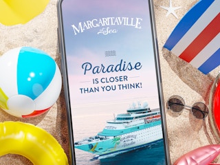A phone sitting on sand with beach accessories around it, the screen displaying an image of the Margaritaville at Sea cruise ship and ...