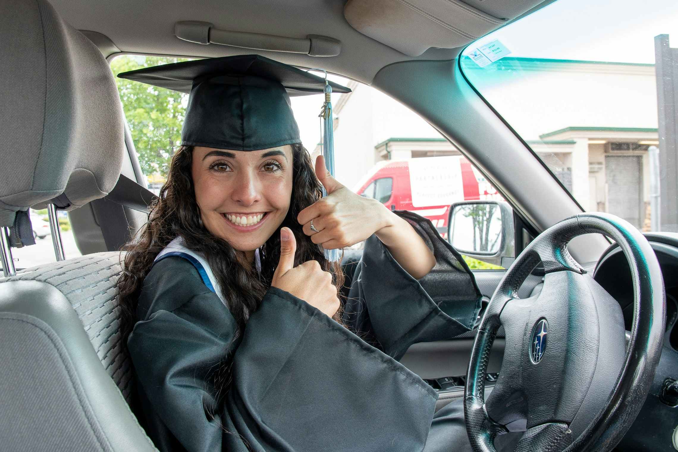 Woman in a graduation cap and gown sitting behind the wheel of a car.