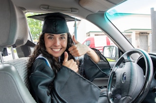 Woman in drivers seat of car, wearing a gradation cap and gown, holding two thumbs up.