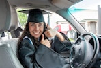 Woman in a graduation cap and gown sitting behind the wheel of a car.