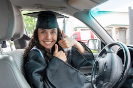 Woman in a graduation cap and gown sitting behind the wheel of a car.