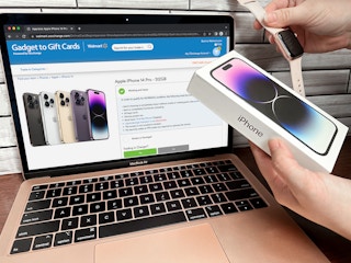 Someone holding an iPhone 14 and Apple Watch in front of a laptop displaying the Walmart electronics trade in webpage