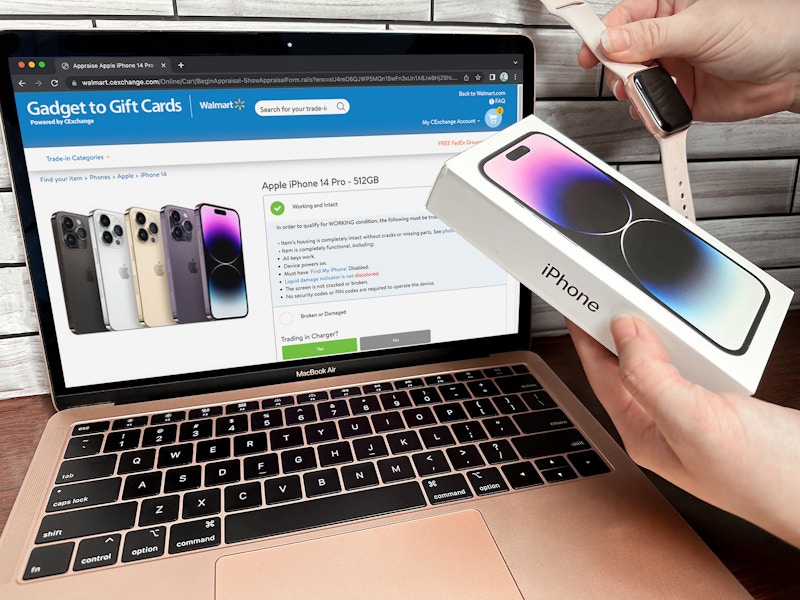 Someone holding an iPhone 14 and Apple Watch in front of a laptop displaying the Walmart electronics trade in webpage