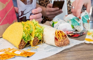 Taco Bell food on a table with people holding drinks and phones in background.