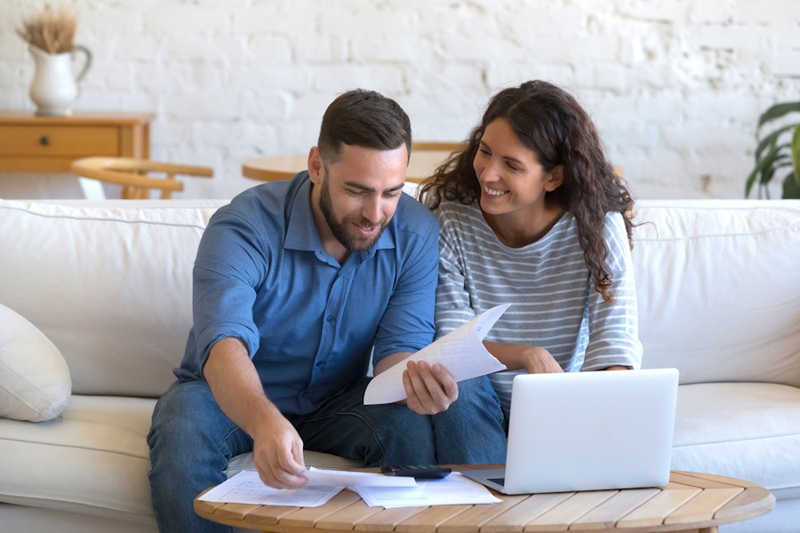 happy couple sitting on sofa looking at paperwork and laptop