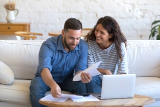 happy couple sitting on sofa looking at paperwork and laptop
