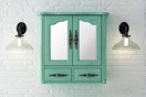 Lifestyle image of a vintage turquoise bathroom vanity mirror cabinet against a white brick wall w/ wall ligths