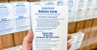 a persons hand holding up a Menards Rebate Form booklet in front of a wall of more booklets.