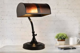Lifestyle image of a brown bankers lamp on a desk with while brick wall in the background