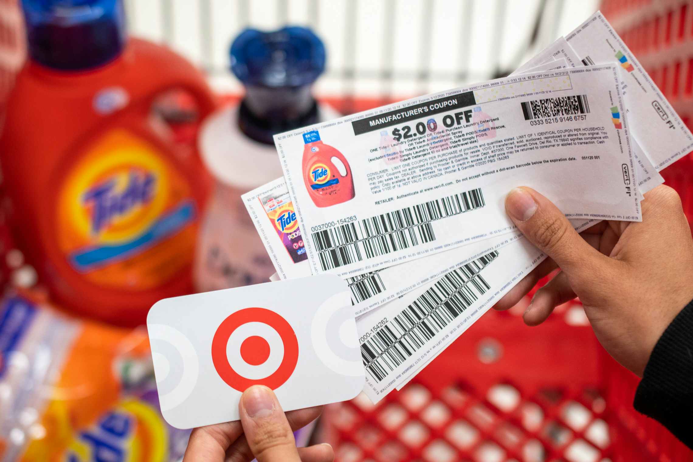 A person holding tide manufacture coupons in one hand and a Target gift card in the other, with a shopping card containing laundry ...