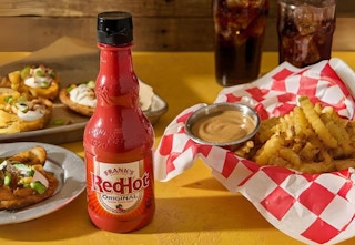 bottle of hot sauce next to lots of fried food and soft drinks on table