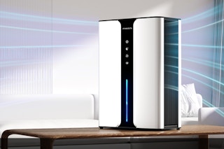 A large white and black dehumidifier sits on a table next to a white couch with a pillow. Blue lines represent moisture leaving the air.