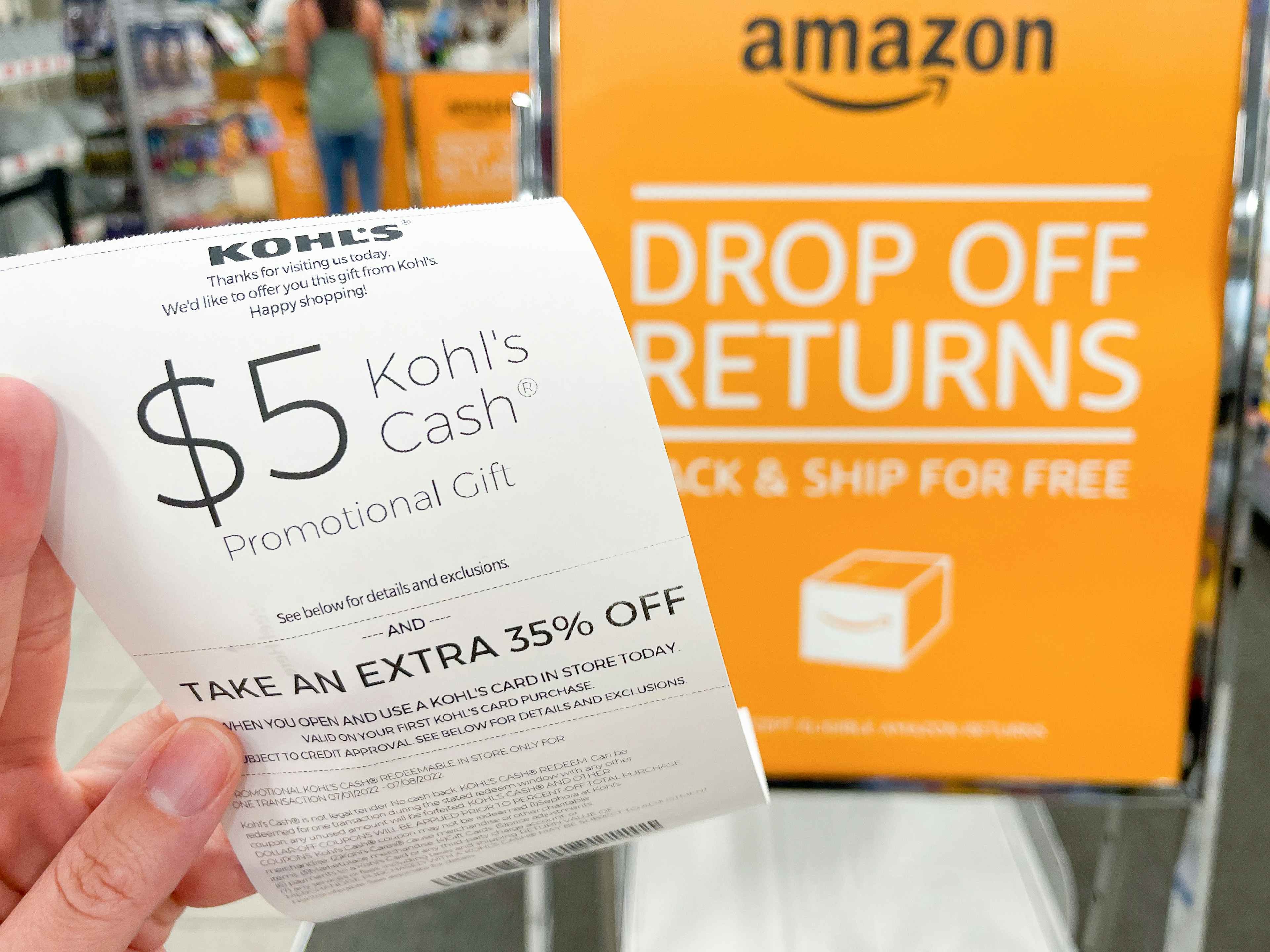A person's hand holding a $5 Kohl's Cash coupon on a receipt for returning an Amazon package next to the Amazon drop off returns sign ins...