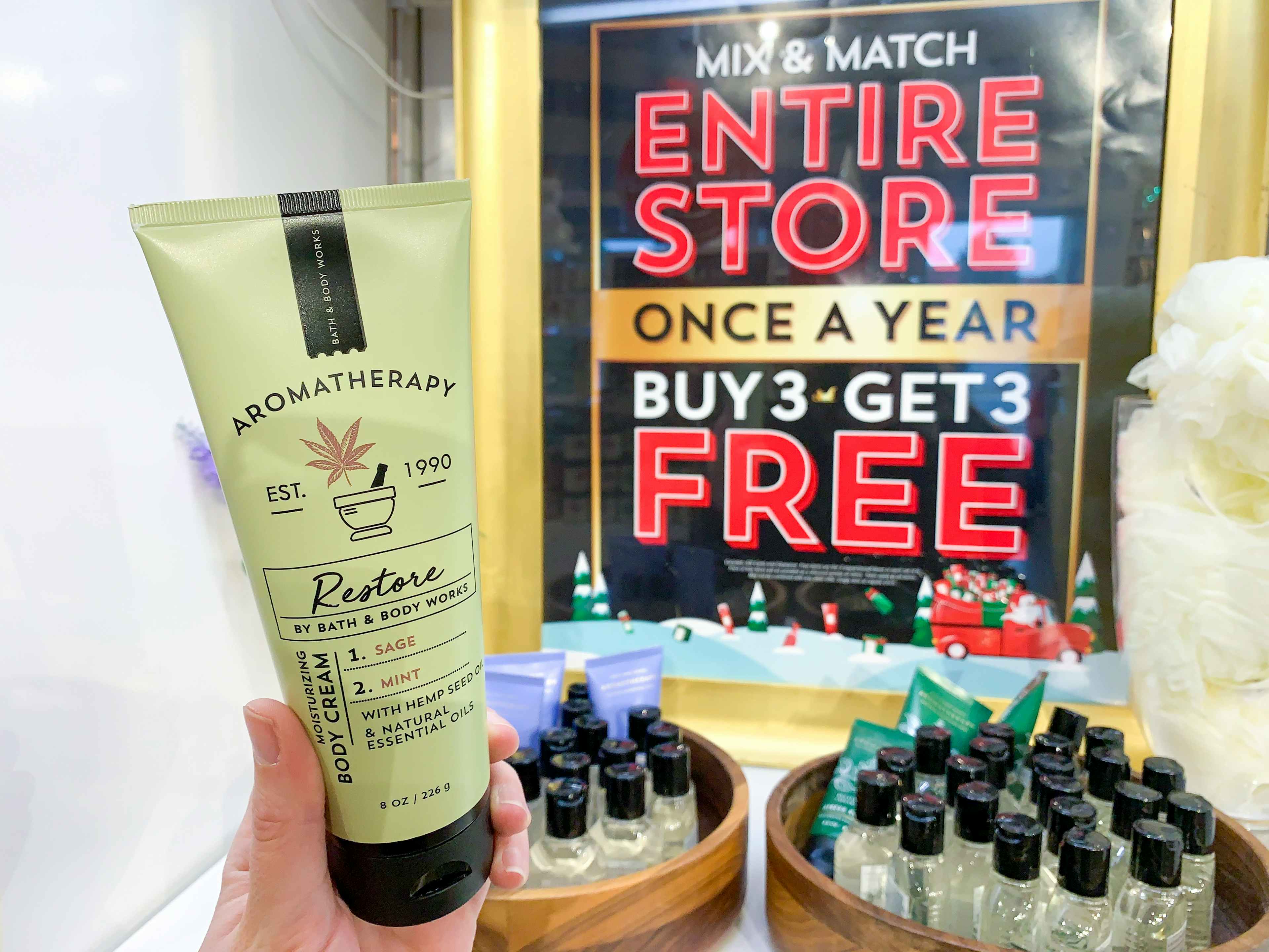 A person's hand holding up a bottle of Aromatherapy lotion in front of a big sign inside Bath & Body Works that reads, "Mix & Match, enti...