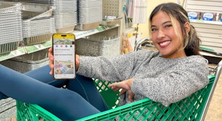 a woman sitting in a dollar tree cart holding a ihpone with the krazy coupon lady app in screen