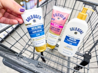 a hand holding a tube of gold bond lotion along with two other bottles of lotion in a shopping cart