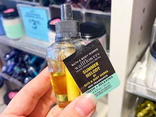 A person's hand holding a Summer Melody wallflower refill in front of the wallflower display inside Bath & Body Works.