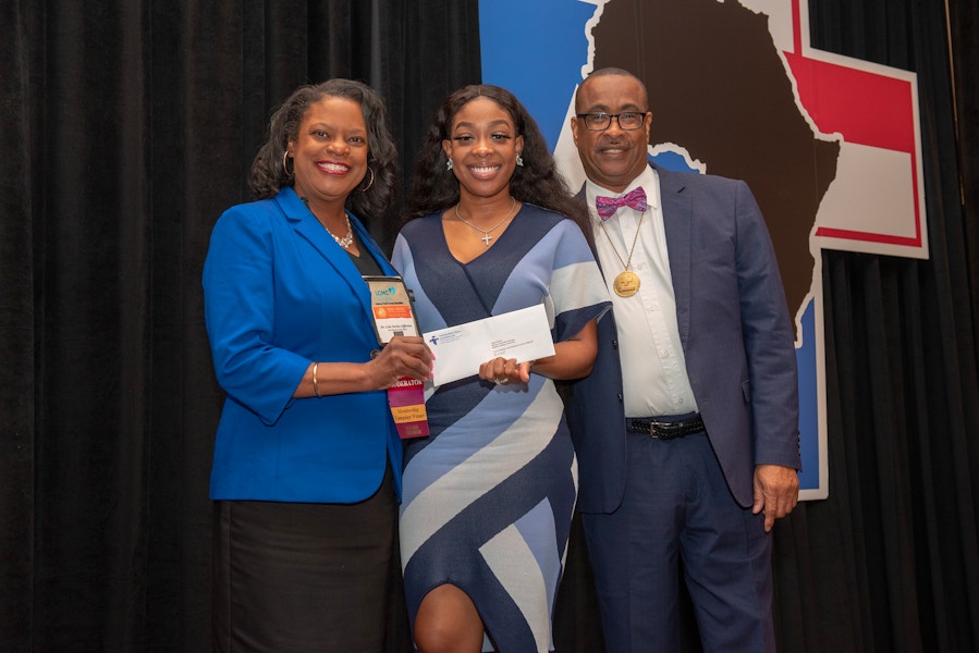 Three people standing on a stage, one of them holding a scholarship envelope.