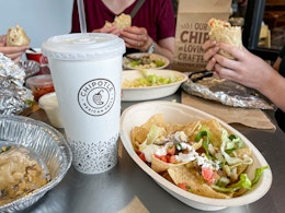 Three people holding burittos at a table filled with food. A drink cup is in the for front clearly displaying the Chipotle logo.
