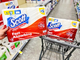 hand holding pack of paper towels in store aisle in front of shopping cart with another pack of paper towels