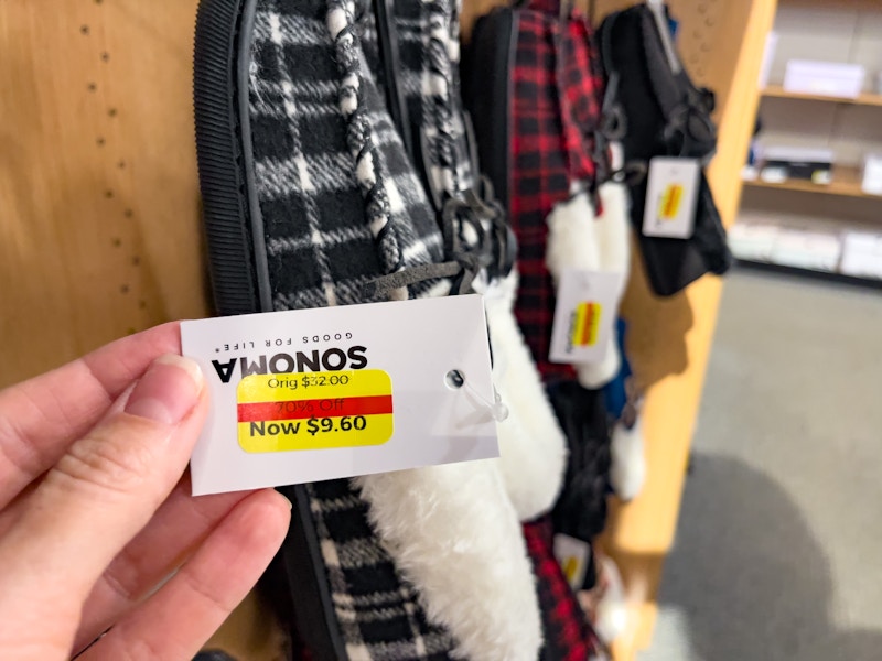 A hand holding a clearance tag hanging from a pair of slippers with other slippers in the background.