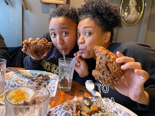 Two women sitting at a table in Cracker Barrel, drinking from the same cup with two different straws, holding up fried chicken.