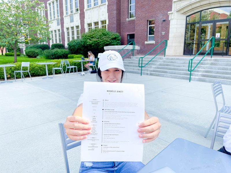 A college student sitting outside of a building on campus and holding a resume up to the camera.