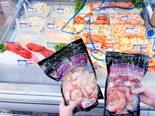 A person's hands holding up two bags of Royal Tiger Wild Ocean Shrimp in front of the seafood display at WinCo Foods.