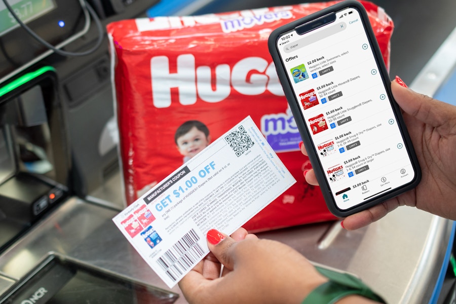 Ibotta app on a cell phone next to coupons and Huggies diapers at a Walmart checkout stand.