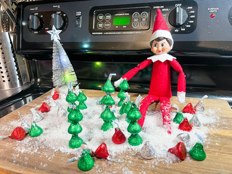 an elf on the shelf doll sitting on a cutting board with fake snow and christmas trees made out of hershey kisses