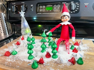 an elf on the shelf doll sitting on a cutting board with fake snow and christmas trees made out of hershey kisses