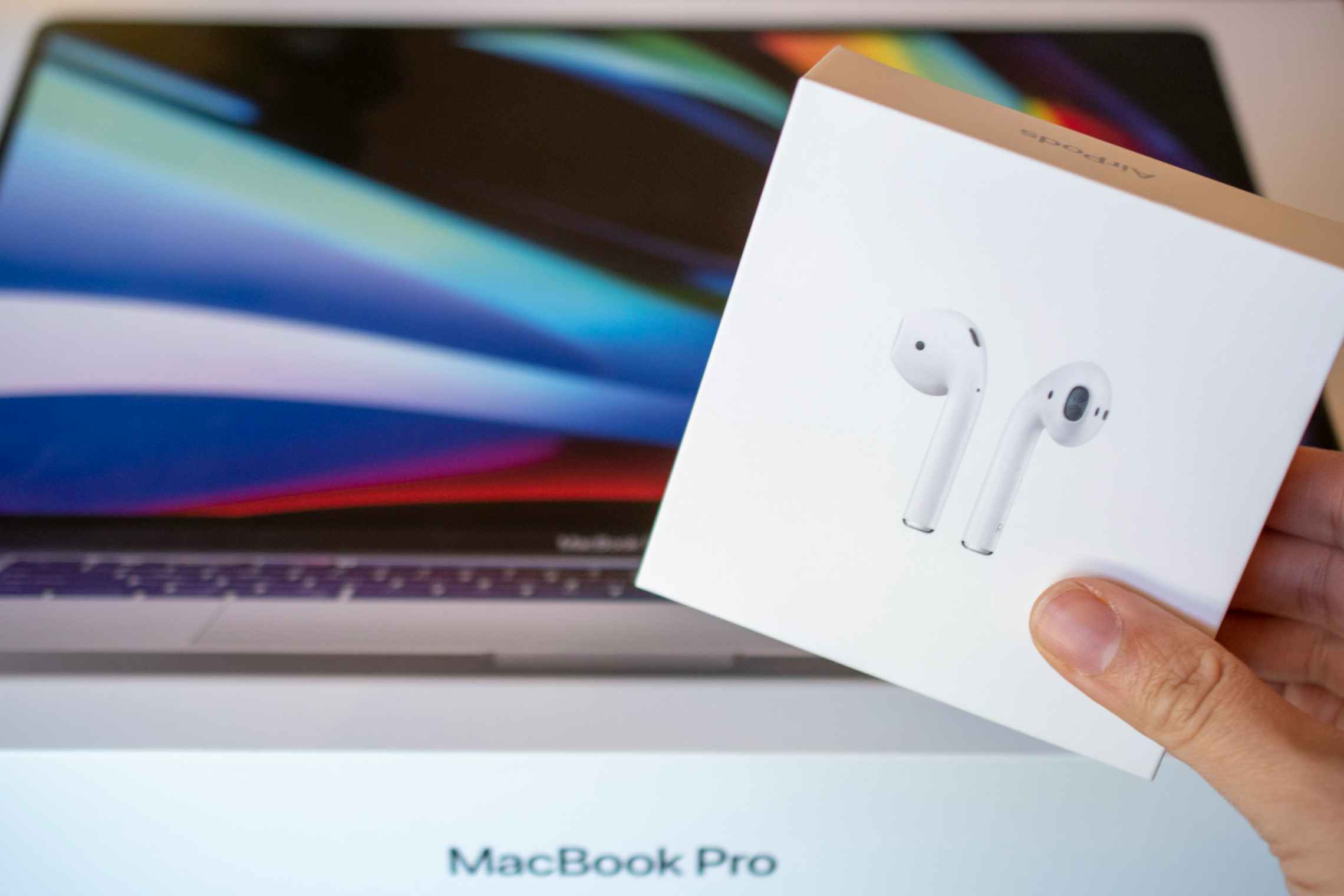 An Apple Airpods box being held in front of a Macbook Pro laptop computer.