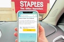 A person holding up a cell phone displaying the Amazon return screen while parked in the parking lot at a Staples store