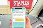 A person holding up a cell phone displaying the Amazon return screen while parked in the parking lot at a Staples store
