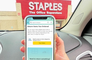 A person holding up a cell phone displaying the Amazon return screen while parked in the parking lot at a Staples store