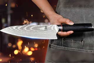 A person in a grey denim apron holds a steel chef knife with a wood handle in front of them. Lights or sparks are behind them.