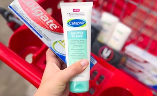 hand holding a bottle of cetaphil acne cream cleanser and colgate over a red target cart