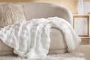 In a beige living room on a beige couch, next to a faux fur beige pillow is a white 50 x 60 fluffy faux fur blanket. Someone should fold it.