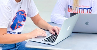 Two college students sitting outside at a table, working on their laptops.
