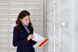 A woman standing at a wall of mail boxes, looking through a handful of envelopes.