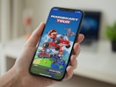 A person's hand holding a phone displaying the loading screen for the Mario Kart Tour online game mobile app.