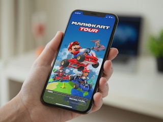 A person's hand holding a phone displaying the loading screen for the Mario Kart Tour online game mobile app.
