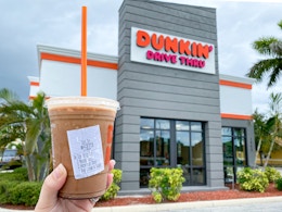 A person's hand holding up a specialty iced coffee outside of a Dunkin restaurant.