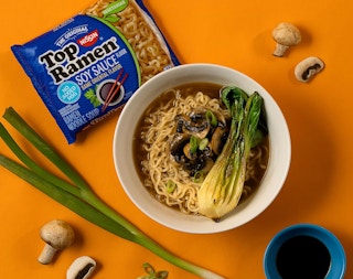 a bowl of ramen next to a pack of ramen and green onions on an orange surface