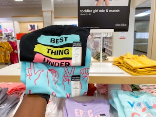 someone holding up two pairs of kids shirts in front of a display of kids clothes