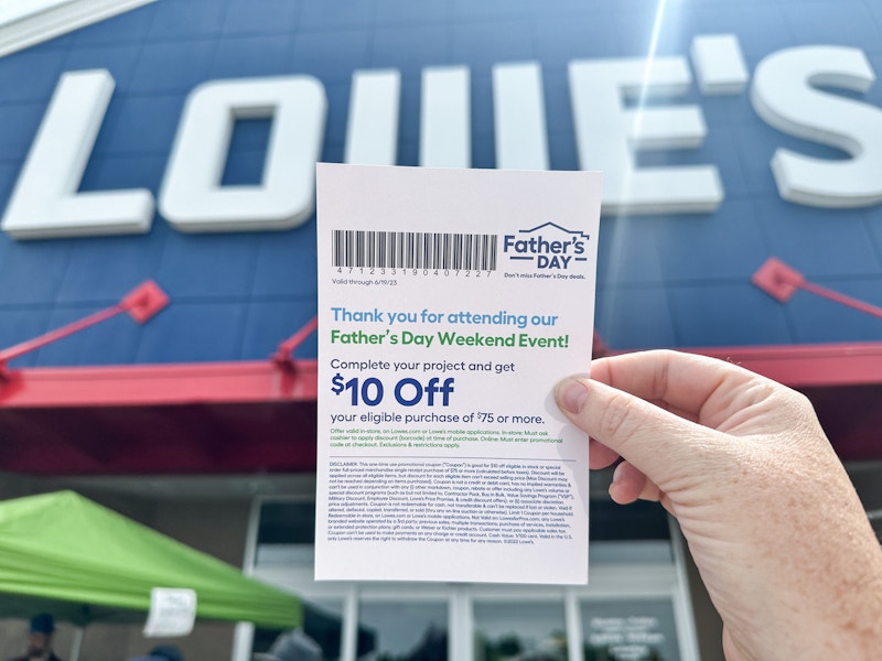 A woman's hand holding up a $10 off coupon in feront of the Lowe's storefront sign