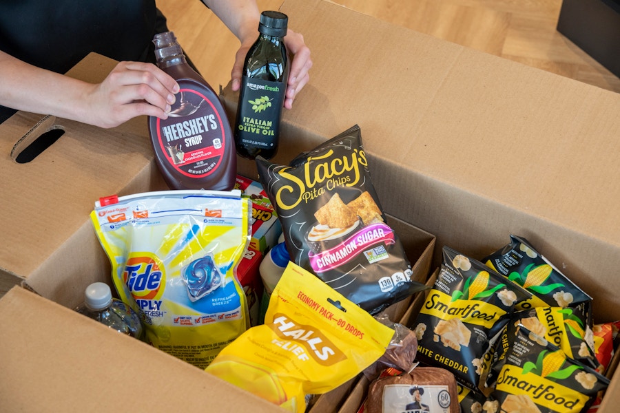 A person's hands holding up Hershey's chocolate syrup and Amazon Fresh Italian Olive Oil above a box of various groceries, including Stac...