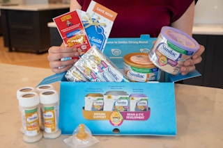 A woman standing in a kitchen, holding products above a Little Gift Similac formula box.